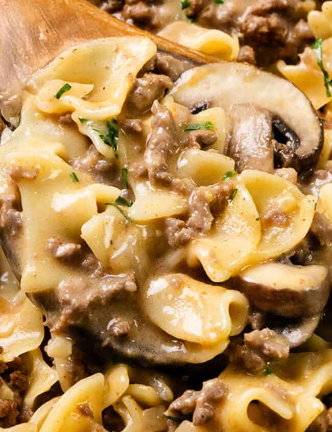 A close-up of an easy beef and mushroom stroganoff with wide egg noodles, garnished with chopped parsley. The creamy sauce coats the ingredients, making it perfect for simple yet satisfying dinners, and a wooden spoon is partially visible.