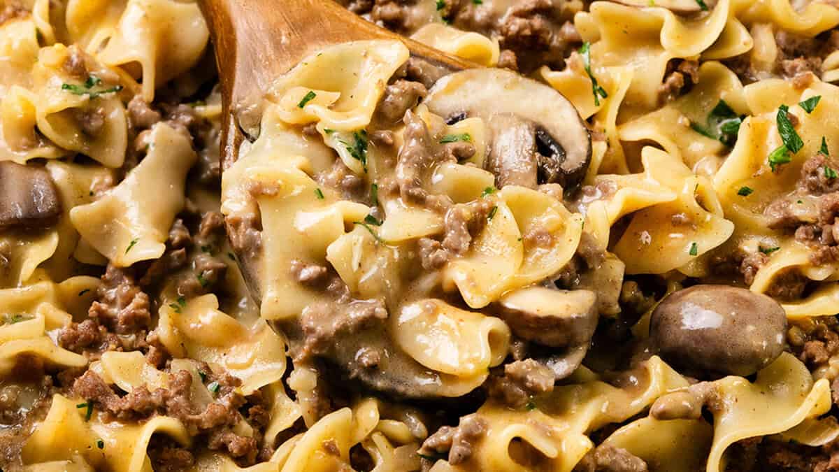 A close-up of an easy beef and mushroom stroganoff with wide egg noodles, garnished with chopped parsley. The creamy sauce coats the ingredients, making it perfect for simple yet satisfying dinners, and a wooden spoon is partially visible.