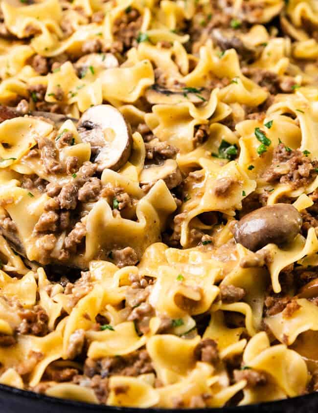 A skillet filled with beef stroganoff features wide egg noodles, ground beef, mushrooms, and a creamy sauce—a true comfort food recipe. Chopped parsley is sprinkled on top, with a wooden spoon resting in the dish, inviting you to dive into its soothing flavors.