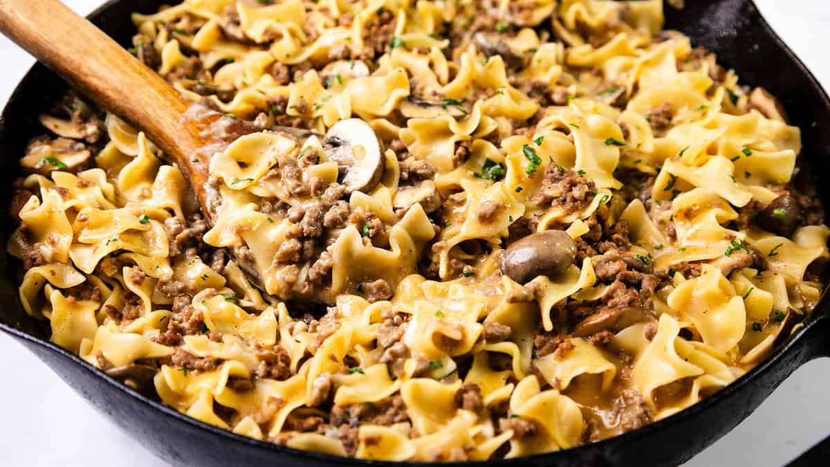 A skillet filled with beef stroganoff features wide egg noodles, ground beef, mushrooms, and a creamy sauce—a true comfort food recipe. Chopped parsley is sprinkled on top, with a wooden spoon resting in the dish, inviting you to dive into its soothing flavors.