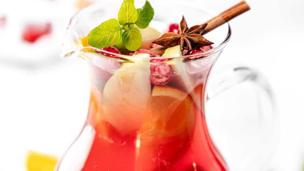 A glass pitcher filled with a vibrant red beverage, garnished with orange slices, cranberries, a cinnamon stick, anise star, and fresh mint leaves against a white background.