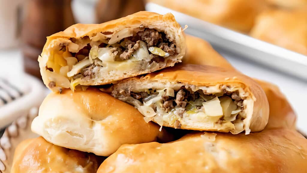 Close-up of freshly baked pastries stuffed with ground meat, cabbage, and onions, displayed in a pile. The flaky golden-brown crust is slightly cracked, revealing the savory filling inside.