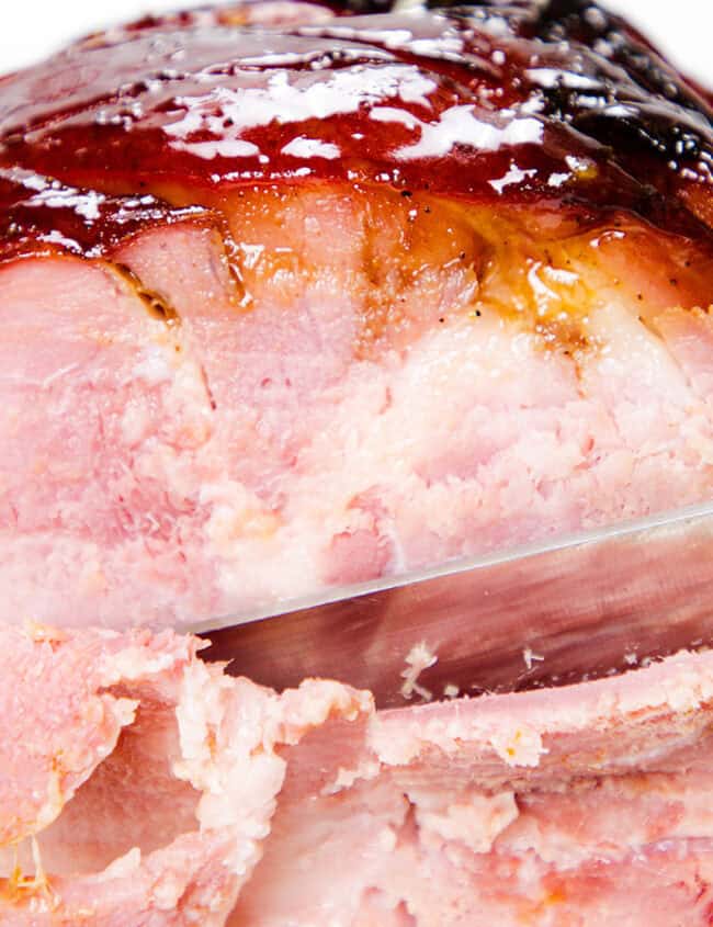 A close-up of a glazed ham being sliced with a serrated knife. The ham has a shiny, caramelized outer layer with a pink, juicy interior. The knife has just cut through a small portion, revealing the tender inside.