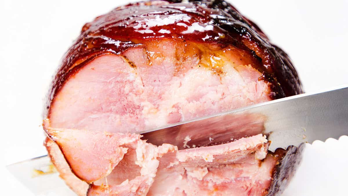 A close-up of a glazed ham being sliced with a serrated knife. The ham has a shiny, caramelized outer layer with a pink, juicy interior. The knife has just cut through a small portion, revealing the tender inside.