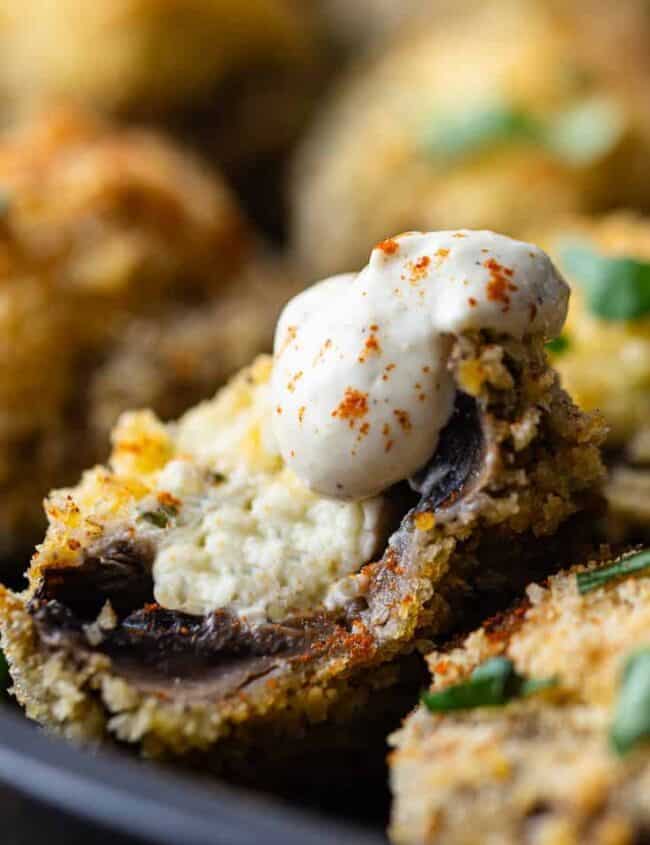 A tantalizing take on mushroom recipes, this close-up reveals stuffed mushrooms topped with a dollop of creamy sauce and sprinkled with paprika. The crispy breadcrumb coating is garnished with fresh herbs, served in a black dish for an irresistibly savory presentation.