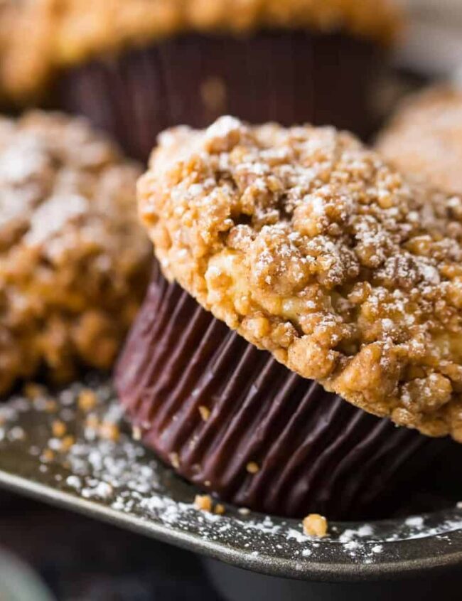 A close-up of freshly baked muffins with a crumbly topping, dusted with powdered sugar, showcases a scene straight out of popular muffin recipes. The muffins rest in a metal pan, with cinnamon sticks slightly visible in the background, creating a warm and inviting atmosphere.