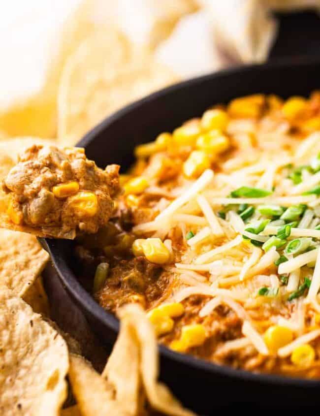 A skillet filled with creamy dip, topped with melted cheese, corn, and chopped green onions. Tortilla chips are arranged around the skillet, with one chip dipping into the mixture.