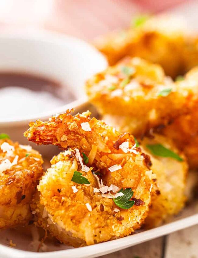 A close-up of a plate with crispy, golden-brown fried shrimp garnished with parsley offers a delicious dinnertime treat. A small bowl of dipping sauce is placed on the side, making this an enticing dinner idea served on a rectangular white plate.