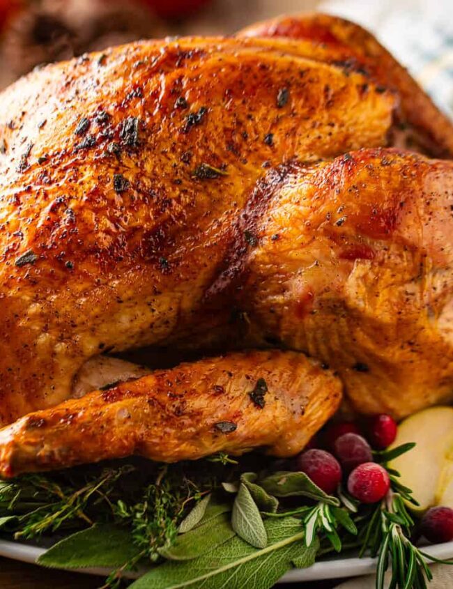 A golden-brown roasted turkey on a white platter garnished with fresh herbs, cranberries, and apple slices. In the background, there's a small pumpkin and a striped napkin, adding a festive touch.