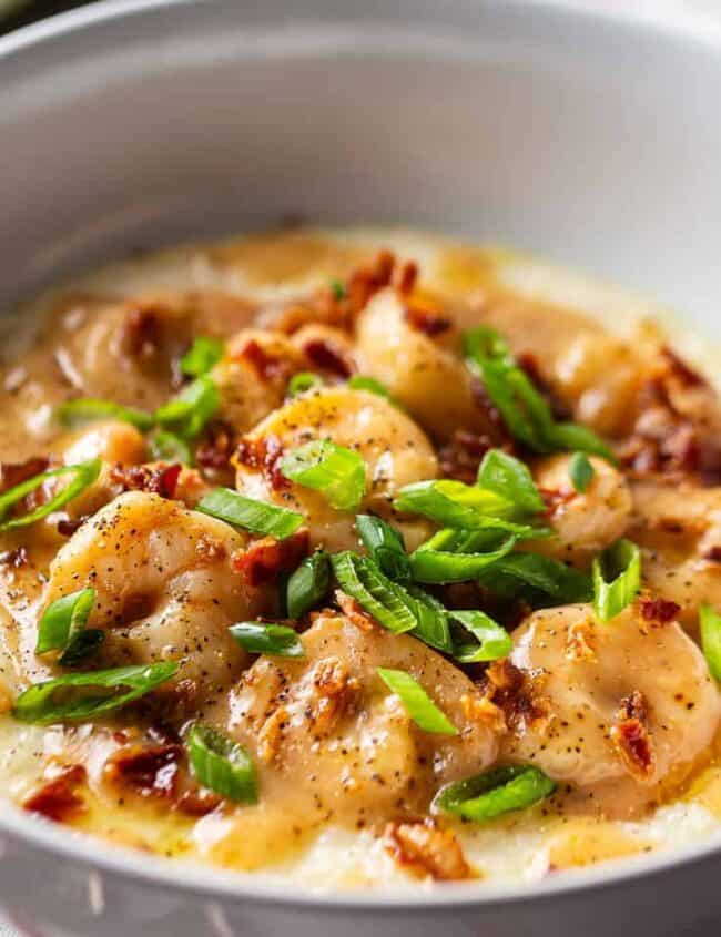 A bowl of creamy grits topped with shrimp, bacon crumbles, and sliced green onions, this delightful seafood recipe is seasoned with black pepper and artfully garnished, making for an appetizing presentation on a wooden table.