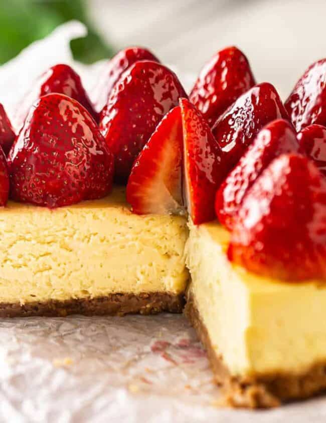 A creamy strawberry dessert cheesecake topped with glossy, fresh strawberries sits on a piece of parchment paper. It has a golden-brown crust, and a slice has been removed, revealing the smooth texture of the cake. Green leaves are blurred in the background.