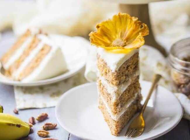 A slice of layered banana cake, inspired by classic cake recipes, boasts white frosting and a yellow flower on top. It rests on a white plate with a fork nearby. In the background, another slice and scattered pecans complete the tempting scene.