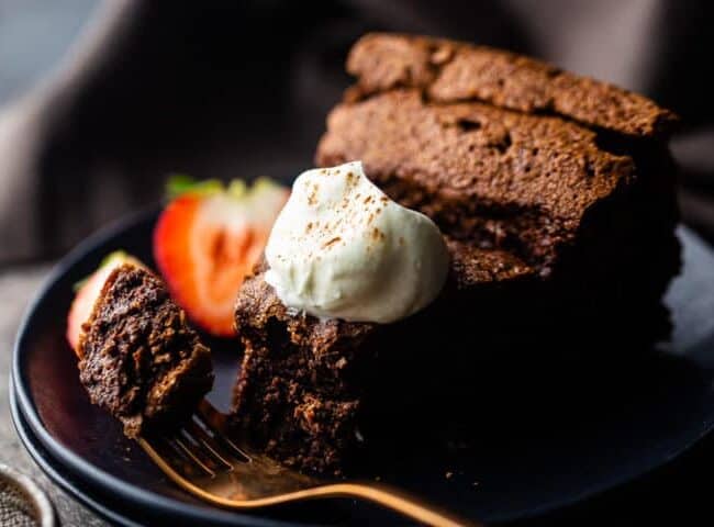 A slice of chocolate cake sits elegantly on a black plate, topped with a dollop of whipped cream. A fork holds a tempting piece, while a strawberry garnish adds a pop of color, inviting you to explore more delicious chocolate recipes.