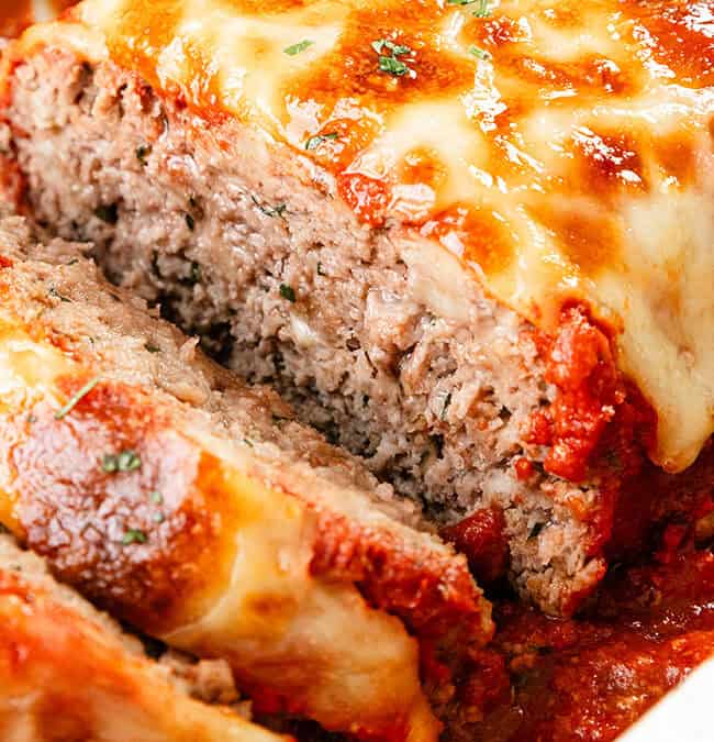 Close-up of a sliced meatloaf made with ground beef, topped with melted cheese and tomato sauce, garnished with herbs in a white dish. Perfect for hearty recipes!