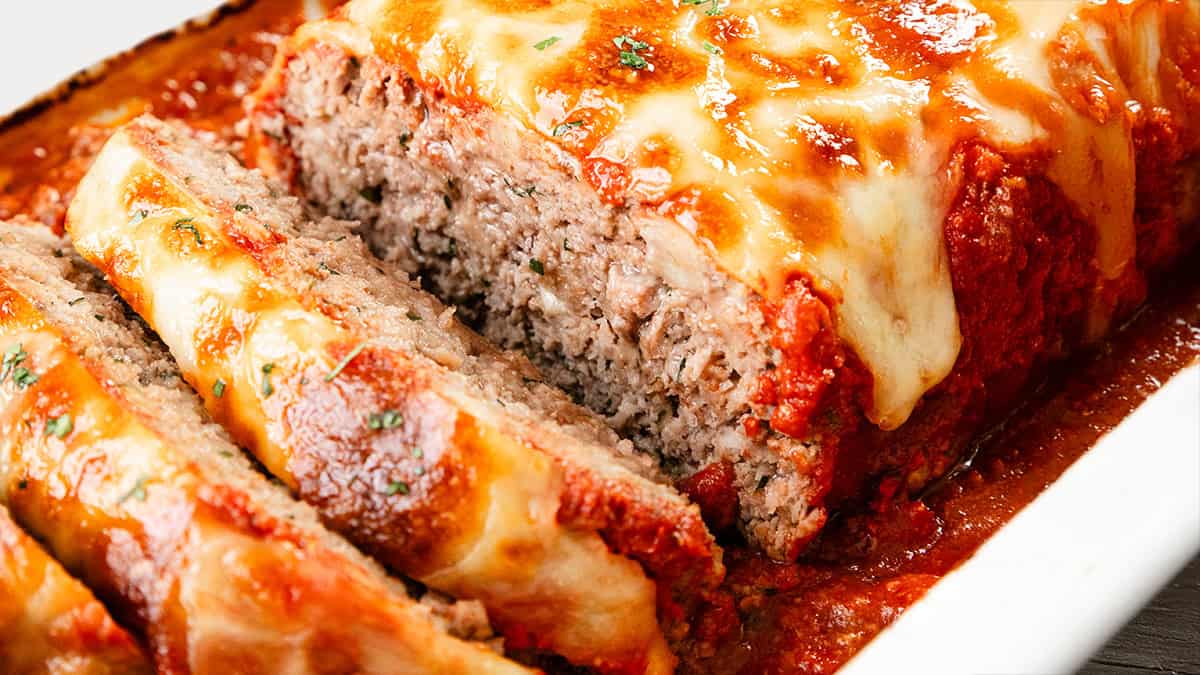 Close-up of a sliced meatloaf made with ground beef, topped with melted cheese and tomato sauce, garnished with herbs in a white dish. Perfect for hearty recipes!