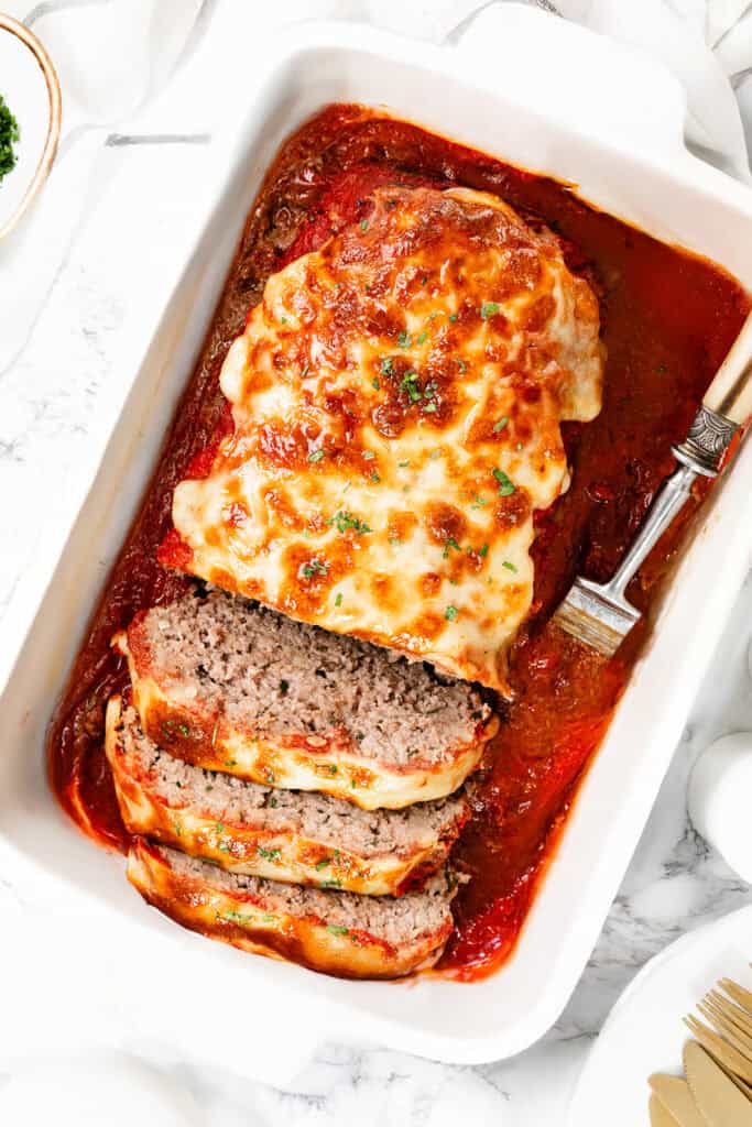 A rectangular white dish showcases an Italian-inspired meatloaf topped with melted cheese and garnished with herbs. Several slices are cut at one end, revealing the juicy interior. A fork rests beside the meatloaf, which is elegantly surrounded by a rich red sauce.