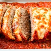 Close-up of a sliced Italian meatloaf in a baking dish, topped with melted cheese and garnished with herbs, resting in a bed of rich tomato sauce.