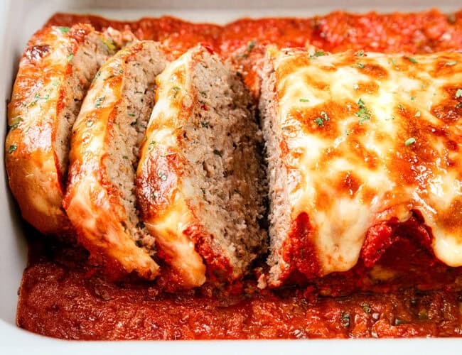 Close-up of a sliced Italian meatloaf in a baking dish, topped with melted cheese and garnished with herbs, resting in a bed of rich tomato sauce.