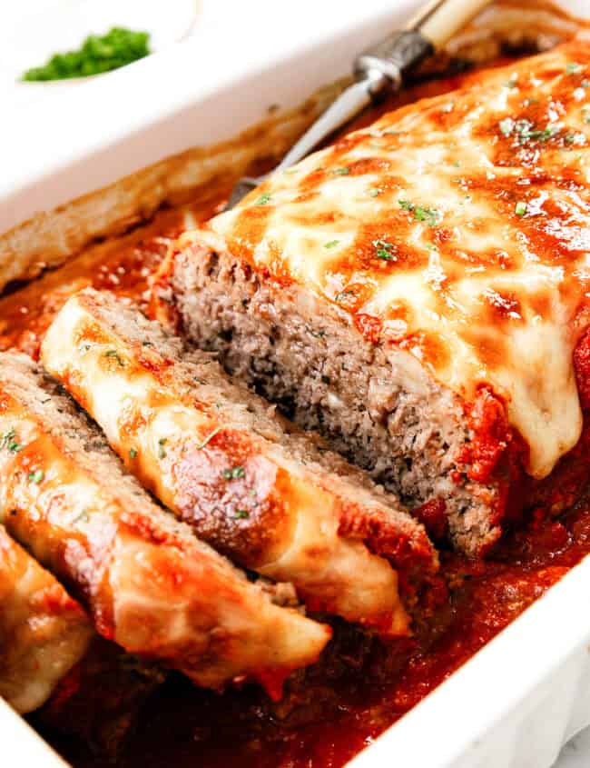 An Italian-inspired meatloaf topped with melted cheese and garnished with herbs is beautifully sliced in a white dish filled with rich tomato sauce. A serving utensil is placed next to it, ready to serve this delicious creation.