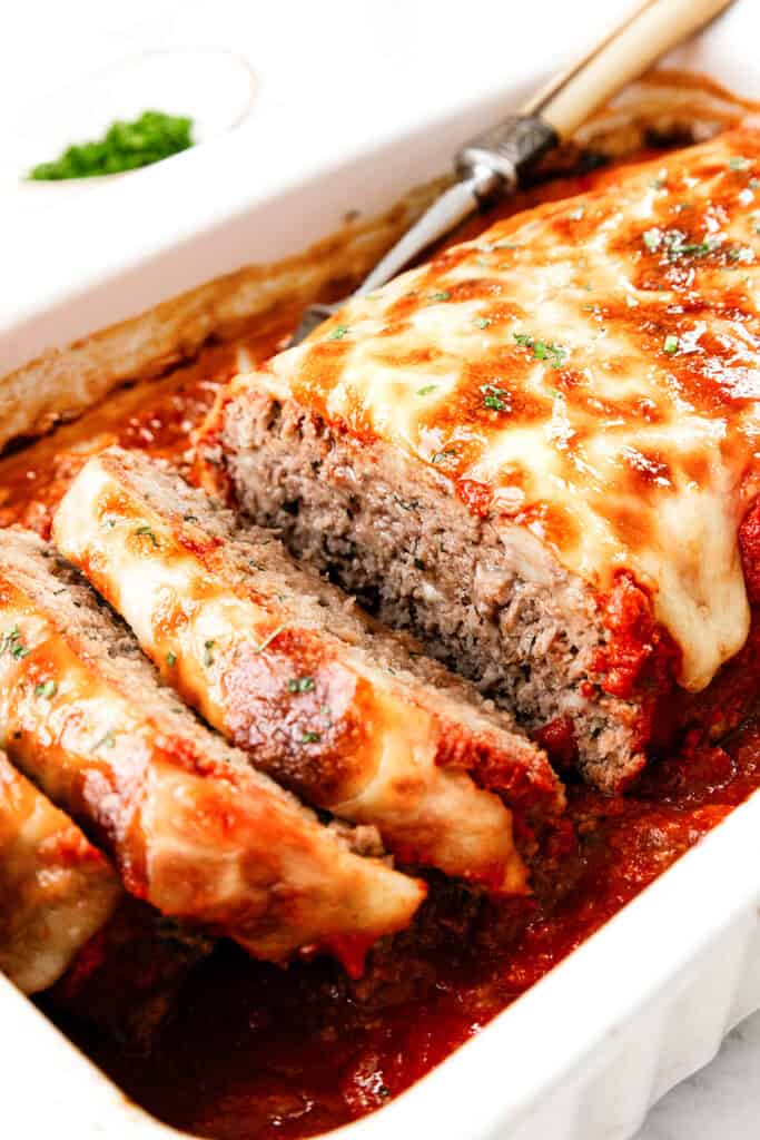 An Italian-inspired meatloaf topped with melted cheese and garnished with herbs is beautifully sliced in a white dish filled with rich tomato sauce. A serving utensil is placed next to it, ready to serve this delicious creation.