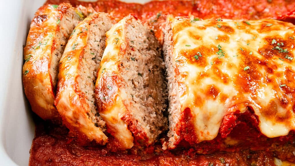 Slices of Italian-inspired cheesy meatloaf topped with melted cheese and tomato sauce in a baking dish. The meatloaf is garnished with herbs, and the sauce appears thick and rich.