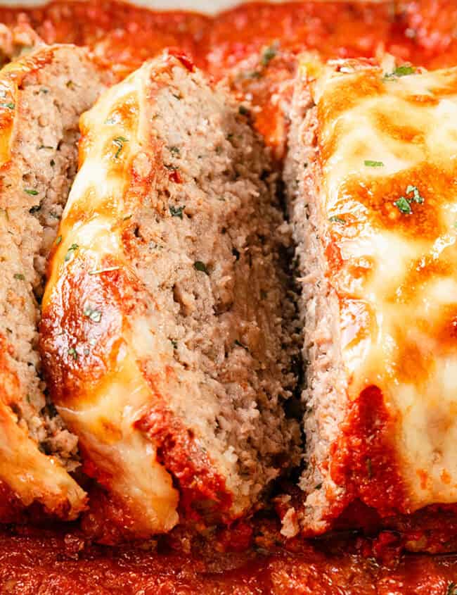 Close-up of a sliced meatloaf topped with melted cheese and herbs, sitting in a pan of rich tomato sauce. This handed down recipe features a golden-brown crust, with each slice lovingly garnished with fresh green herbs.