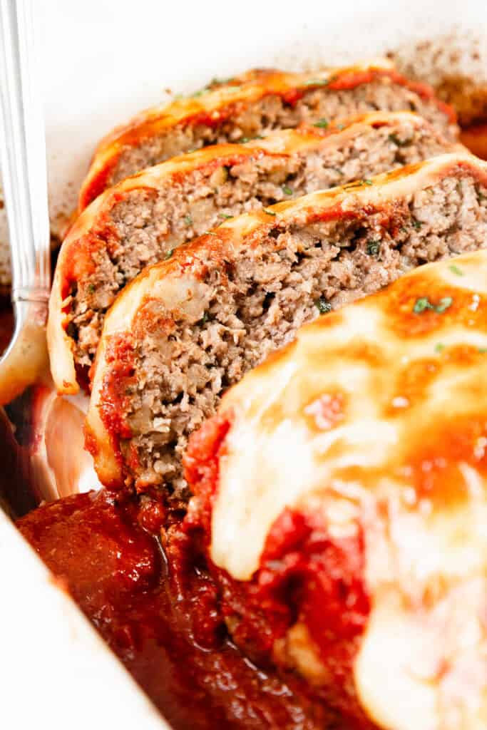 Close-up of sliced Italian meatloaf topped with melted cheese and tomato sauce in a baking dish. A silver serving spoon is partially visible on the side. The seasoned meatloaf, rich with Italian cuisine flavors, is browned with juices pooling at the bottom of the dish.