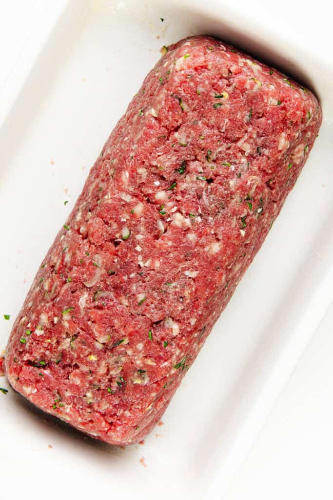 A rectangular block of Italian-style meatloaf, infused with herbs, is displayed on a white surface. The mixture has a reddish tint with visible green and white flecks, showcasing the coarse texture typical of ground meat preparations.