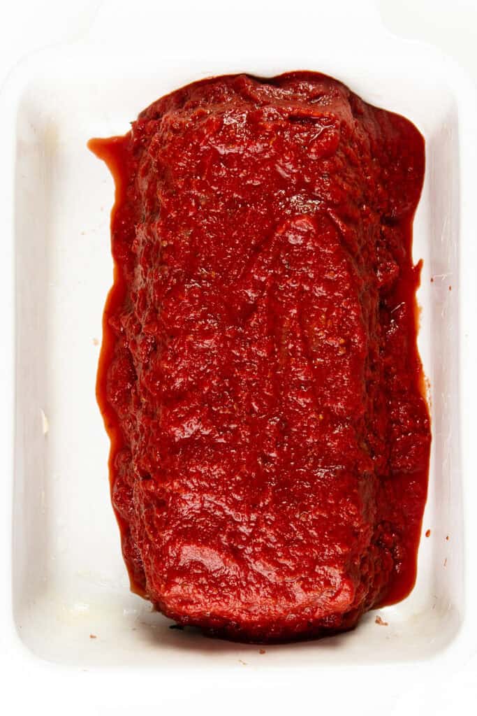 An Italian-inspired meatloaf coated with a rich, red tomato-based sauce is placed in a rectangular white baking dish. The meatloaf is centered and surrounded by a flavorful layer of sauce.