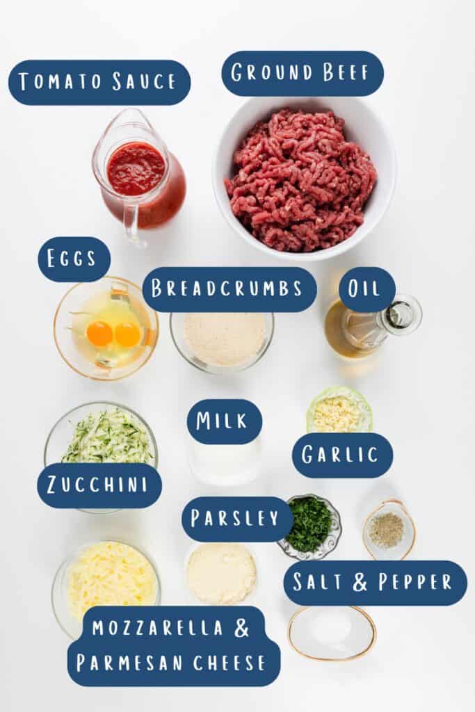 A flat lay of Italian-inspired ingredients labeled for a meatloaf recipe: tomato sauce, ground beef, eggs, breadcrumbs, oil, zucchini, milk, parsley, garlic, mozzarella & parmesan cheese, salt & pepper in bowls and containers on a white background.