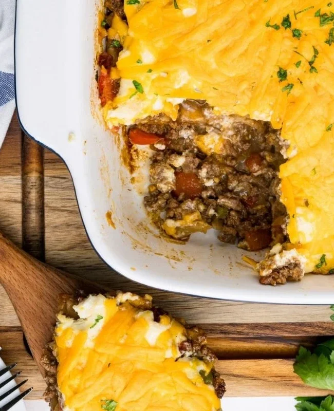 A dish of shepherd's pie with a slice cut out, revealing layers of ground meat, vegetables, and fluffy mashed potatoes, topped with melted cheese. A wooden serving spoon is placed nearby, and fresh parsley is on the side—a true classic from comfort food recipes.