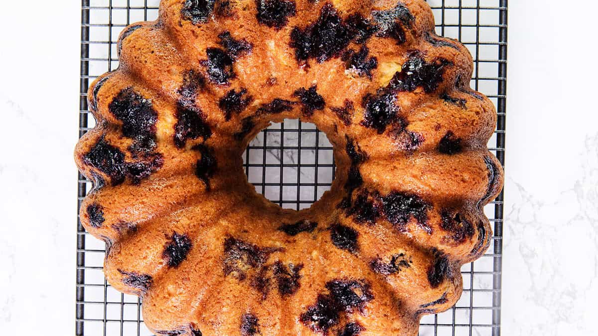 A freshly baked bundt cake with a swirl of dark berry compote sits on a cooling rack. The cake has a golden brown crust with deep purple spots from the berries, creating a marbled effect. The cooling rack rests on a white surface.