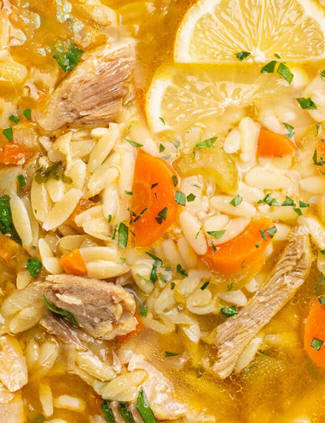 A comfort bowl of soup featuring orzo pasta, shredded chicken, and sliced carrots, garnished with fresh parsley and lemon wedges. A spoon rests on the side, and the dish is placed on a white marble surface.