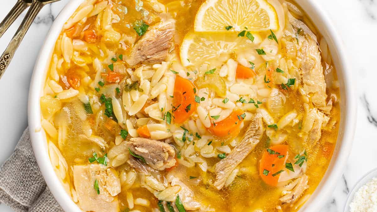 A comfort bowl of soup featuring orzo pasta, shredded chicken, and sliced carrots, garnished with fresh parsley and lemon wedges. A spoon rests on the side, and the dish is placed on a white marble surface.