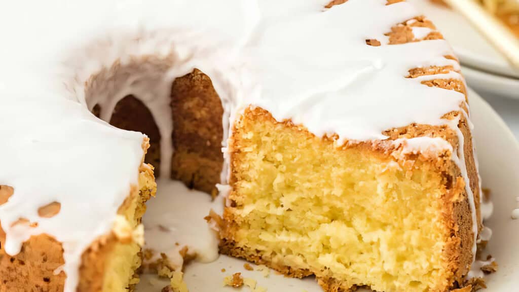 A close-up of a sliced Bundt cake highlights this delectable dessert with a lemon glaze. The cake is golden brown, featuring a moist, dense crumb as the white glaze drips enticingly down its sides onto the plate. A small slice is missing, revealing its irresistible texture.