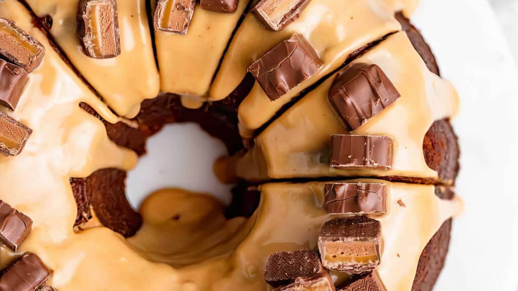 A close-up of a decadent chocolate bundt cake topped with creamy caramel glaze and decorated with chunks of chocolate bars. This dessert is sliced, revealing its moist texture underneath the glossy coating, making it an irresistible delight for any baking enthusiast.