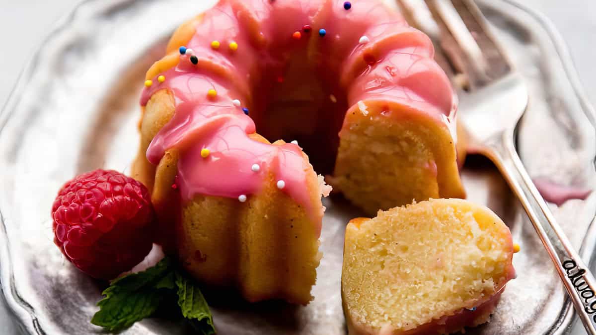 A delectable Bundt cake with pink icing and colorful sprinkles sits on a silver plate. A slice is cut to reveal its moist interior, showcasing the art of baking. Nearby, a raspberry and mint leaves add a fresh touch, with a silver fork poised to enjoy the dessert's delightful flavors.