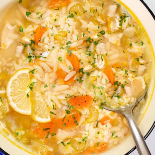 A close-up of a pot filled with lemon orzo soup. Visible ingredients include sliced carrots, celery, chopped parsley, orzo pasta, and a lemon wedge for garnish. The pot has a blue rim, and a metal ladle rests inside.