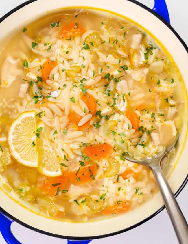 A close-up of a pot filled with lemon orzo soup. Visible ingredients include sliced carrots, celery, chopped parsley, orzo pasta, and a lemon wedge for garnish. The pot has a blue rim, and a metal ladle rests inside.