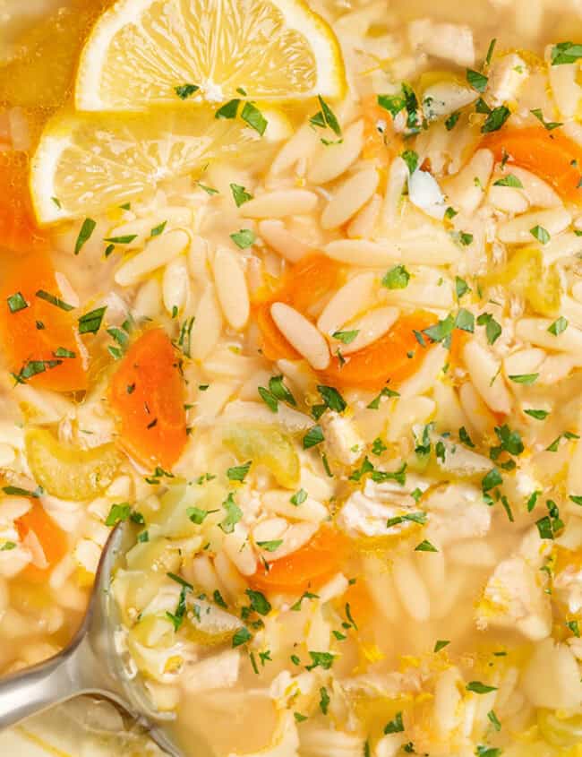 A pot of chicken orzo soup with slices of carrots, celery, lemon wedges, and chopped parsley beckons from within its vibrant blue-edged embrace. Perfect for comfort food recipes, this soup cradles a ladle ready to serve warmth and flavor.