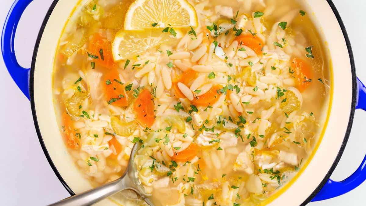 A pot of chicken orzo soup with slices of carrots, celery, lemon wedges, and chopped parsley beckons from within its vibrant blue-edged embrace. Perfect for comfort food recipes, this soup cradles a ladle ready to serve warmth and flavor.