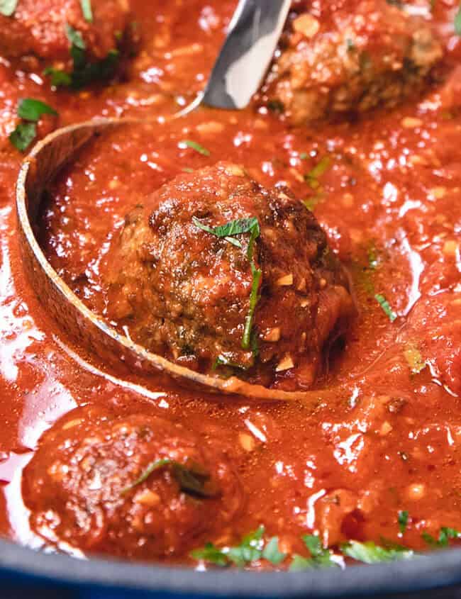 A pot of meatballs covered in rich tomato sauce, garnished with chopped herbs. A wooden spoon is stirring the dish, which is steaming hot and ready to serve.