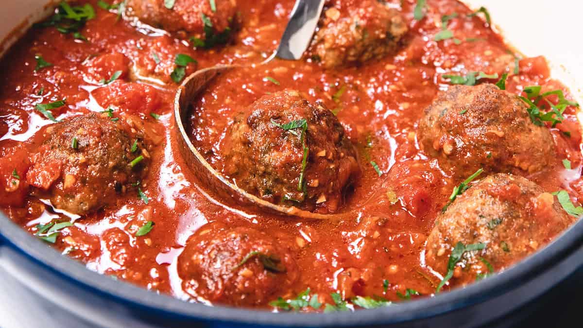 A pot of meatballs covered in rich tomato sauce, garnished with chopped herbs. A wooden spoon is stirring the dish, which is steaming hot and ready to serve.