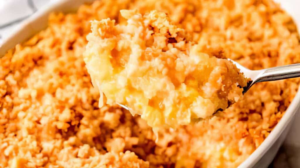 A spoon lifts a serving of cheesy, golden-brown potato casserole with a crispy topping from a baking dish. Creamy potatoes spill out beneath the crunchy, textured crust. I'm sorry, but more details are needed to identify specific SEO keywords for this delicious comfort food scene.