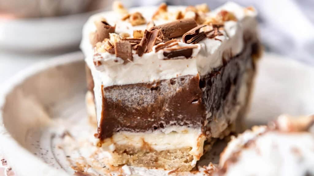 A close-up of a Southern layered dessert slice on a plate. The treat features a crumbly base, creamy white layer, rich chocolate layer, and is topped with whipped cream and chocolate shavings.