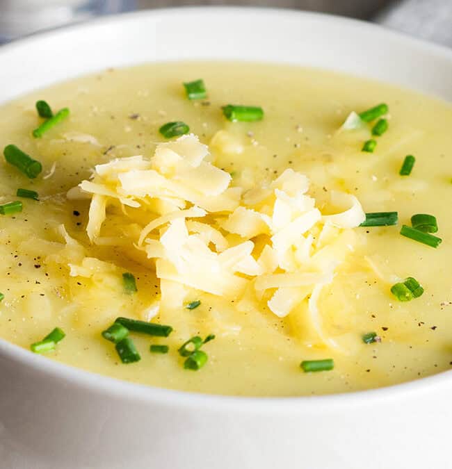 A white bowl filled with creamy potato soup, a staple of old school recipes, topped with shredded cheese and garnished with chopped green onions and ground black pepper. A spoon rests nearby on a napkin.