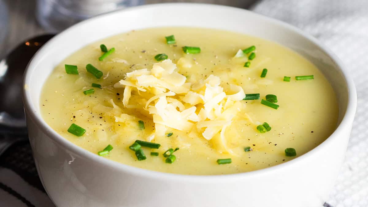 A white bowl filled with creamy potato soup, a staple of old school recipes, topped with shredded cheese and garnished with chopped green onions and ground black pepper. A spoon rests nearby on a napkin.