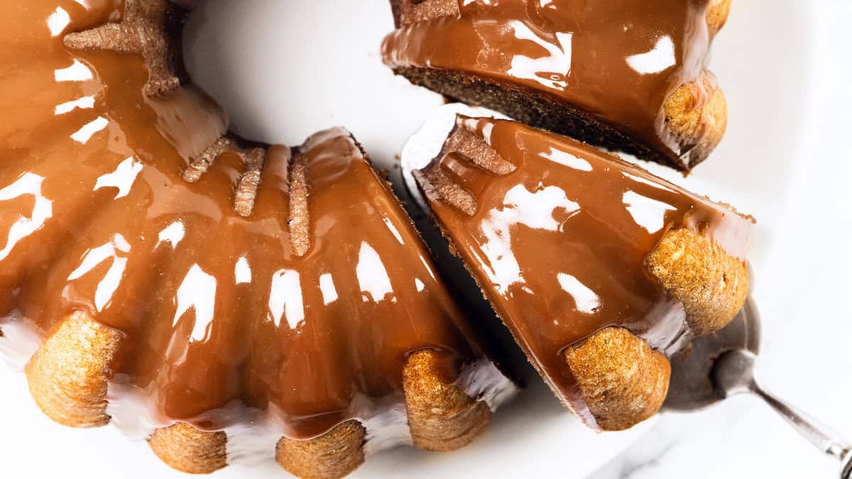 A bundt cake covered in glossy chocolate glaze, with a slice being lifted out on a spatula. The cake is on a white plate and the glaze appears smooth and rich.