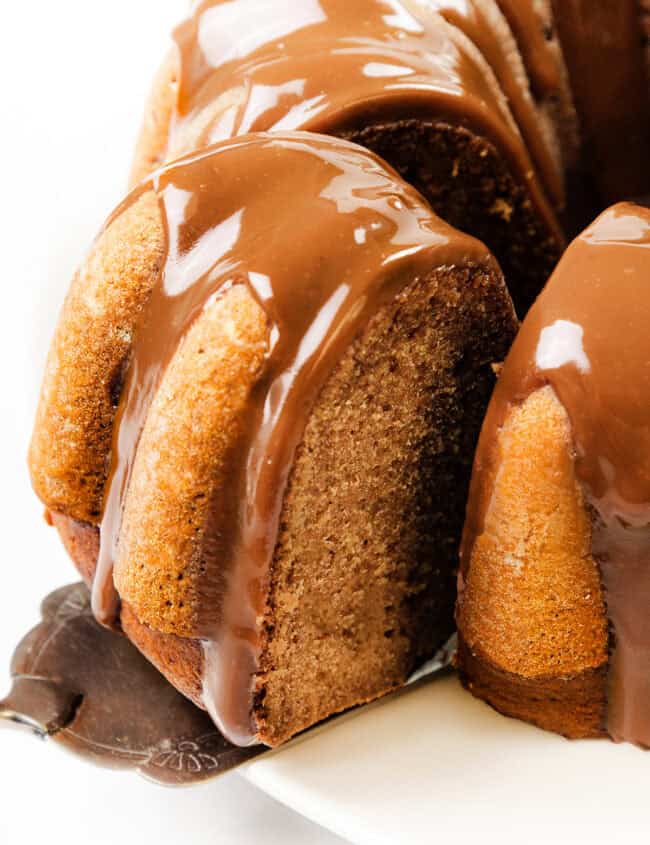 A close-up of a pumpkin Bundt cake on a white plate. The cake is topped with a glossy chocolate glaze that drips down the sides, with one slice partially removed to reveal the moist texture inside.