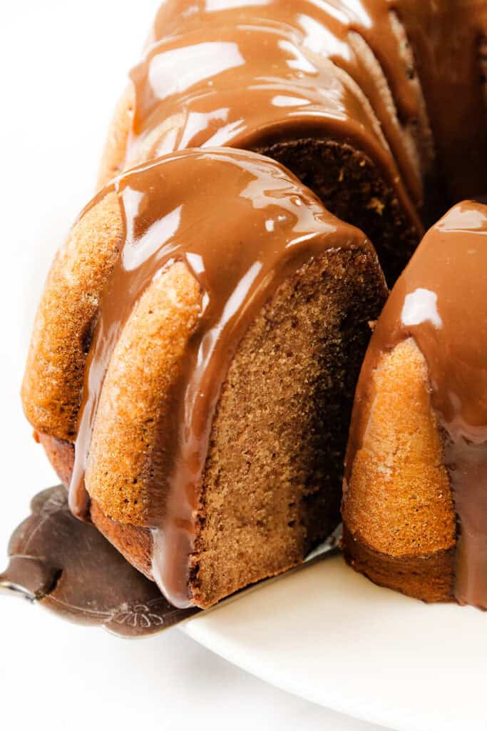 A close-up of a pumpkin Bundt cake on a white plate. The cake is topped with a glossy chocolate glaze that drips down the sides, with one slice partially removed to reveal the moist texture inside.